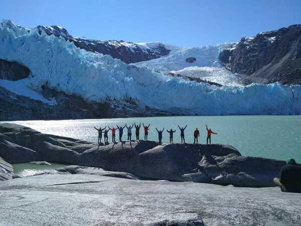 Students standing with their arms up, a bay and a glacier flowing down from mountains are in the background.