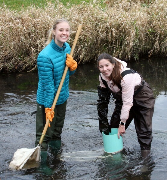 Two women wearing waders stand in the middle of a creek. One is holding a bucket in the water, the other is holding a canvas sample net in the water.